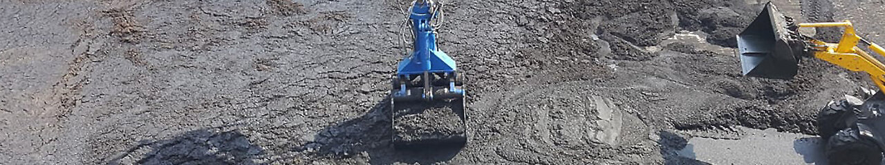 Ansicht von oben, Vogelperspektive, eine Tanks mit schlammartigem Inhalt, welcher durch einen kleinen gelben Bagger, rechts im Bild, zusammen geschoben wird. Ein königsblauer Greifarm, in der Mitte des Bildes, heb beziehungsweise lädt die schlammartige Masse mit seinem Greifer auf..