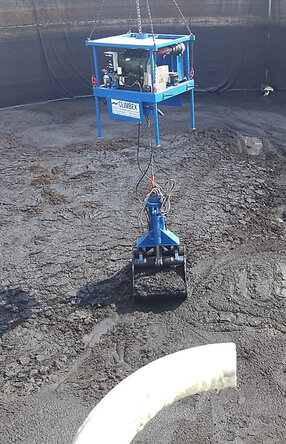 Ansicht von oben, Vogelperspektive, eine Tanks mit schlammartigem Inhalt, welcher durch einen kleinen gelben Bagger, rechts im Bild, zusammen geschoben wird. Ein königsblauer Greifarm, in der Mitte des Bildes, heb beziehungsweise lädt die schlammartige Masse mit seinem Greifer auf..