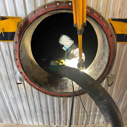 Das Foto zeigt einen von außen Fotografierten Tank mit einer runden Öffnung, im inneren des Tanks steht ein Arbeiter mit Schutzanzug und Maske während er ein Rohr, das von außen kommt, festhält.