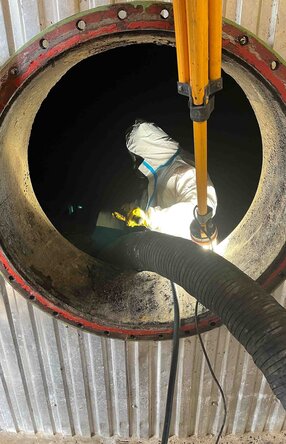 Das Foto zeigt einen von außen Fotografierten Tank mit einer runden Öffnung, im inneren des Tanks steht ein Arbeiter mit Schutzanzug und Maske während er ein Rohr, das von außen kommt, festhält.