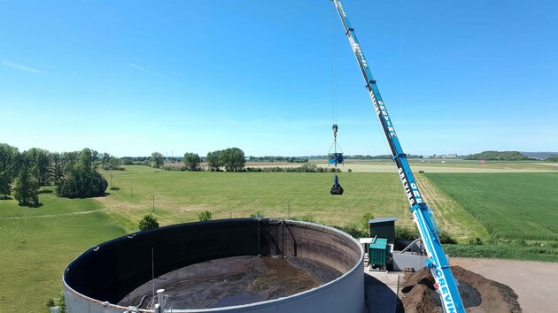 Ein Mitarbeiter sitzt mit Schutzanzug, Gasmaske & Handschuhen auf einem Mini-Bagger der vorne einen Aufsatz zum zusammenschieben von Materalien hat. Er arbeitet im inneren eines Tanks und schieb eine Schlamm ähnliche Substanz rum.