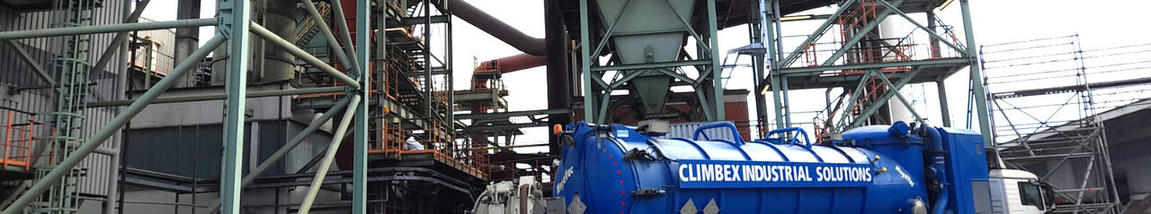 Zu sehen ist das Äußere eines Fabrikgeländes mit diversen Rohren und Metallgeländern, außerdem steht darin eine trichterförmige Anlage. Vor der Anlage steht ein LKW mit einem großen Tank.