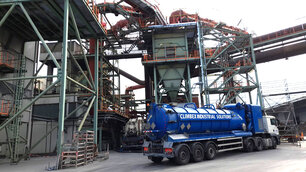 Zu sehen ist das Äußere eines Fabrikgeländes mit diversen Rohren und Metallgeländern, außerdem steht darin eine trichterförmige Anlage. Vor der Anlage steht ein LKW mit einem großen Tank.