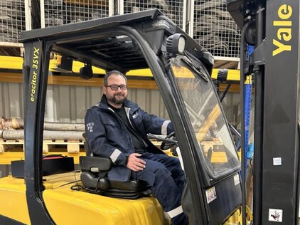 Frontale Aufnahme eines Mannes der in einem Gabelstapler sitzt, er guck leicht zur linken Seite gelehnt in die Kamera. Er ist mittleren Alters, hat einen kurzen grauer Bart sowie Haare, eine Brille und dunkelblauem Ganzkörperanzug.