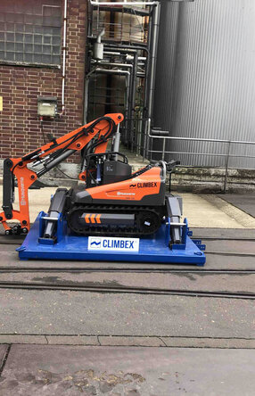 Roboter, orangefarben, mit gesenktem Greifarm stehend auf einer königsblauen Platte die über ein Schienensystem auf einem Betriebshof fährt. Im Hintergrund sind eine Backstein-Fassade und Silos oder Tanks zu sehen.