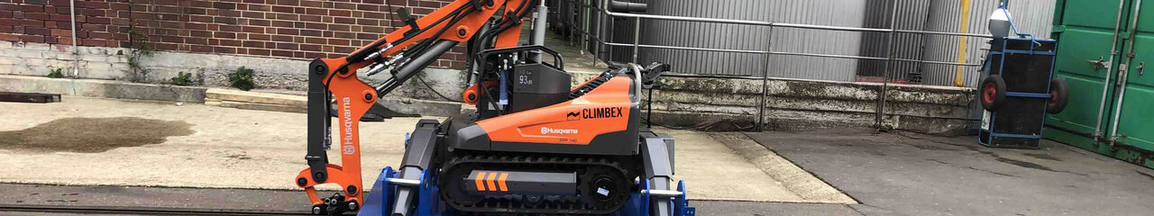 Roboter, orangefarben, mit gesenktem Greifarm stehend auf einer königsblauen Platte die über ein Schienensystem auf einem Betriebshof fährt. Im Hintergrund sind eine Backstein-Fassade und Silos oder Tanks zu sehen.