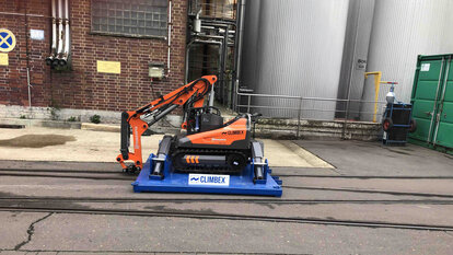 Roboter, orangefarben, mit gesenktem Greifarm stehend auf einer königsblauen Platte die über ein Schienensystem auf einem Betriebshof fährt. Im Hintergrund sind eine Backstein-Fassade und Silos oder Tanks zu sehen.