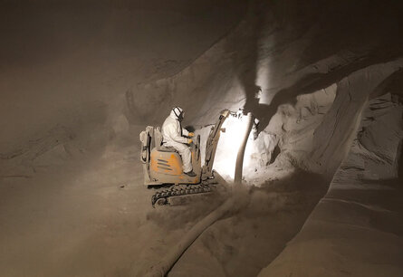 Zu sehen ist ein Arbeiter in Schutzanzug im inneren eines Silos auf einem Bagger ähnliche Fahrzeug an dem ein Schlauch befestigt ist, womit ein Pulver aufgesaugt wird.