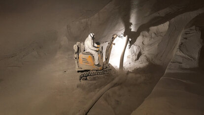 Zu sehen ist ein Arbeiter in Schutzanzug im inneren eines Silos auf einem Bagger ähnliche Fahrzeug an dem ein Schlauch befestigt ist, womit ein Pulver aufgesaugt wird.