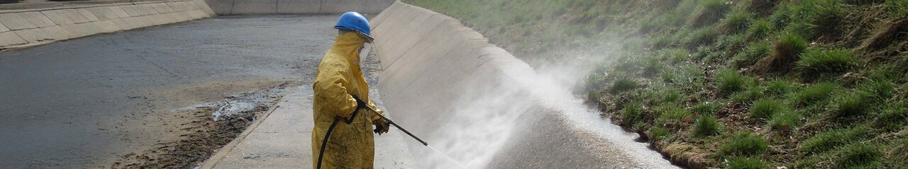 Es steh eine Person mit gelbem Ganzkörperanzug und königsblauen Arbeitshelm in einer offenen, Kanal ähnlichen, Grube und hält dabei einen Hochdruckstrahler auf die Innenwand.