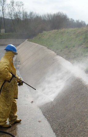 Es steh eine Person mit gelbem Ganzkörperanzug und königsblauen Arbeitshelm in einer offenen, Kanal ähnlichen, Grube und hält dabei einen Hochdruckstrahler auf die Innenwand.