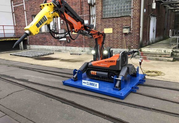 Roboter, orangefarben, mit ausgestrecktem Greifarm, Stift ähnlichem Aufsatz am Ende des Greifarms und kleinen Scheinwerfern, auf einer königsblauen Platte, die über ein Schienensystem auf einem Betriebshof fährt. Im Hintergrund ist eine Backstein-Fassade zu sehen.