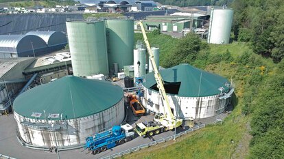 Aufnahme aus der Vogelperspektive, eines grünlichen Fabrikgebäudes, welches aus zwei großen flachen Silos im Vordergrund und zwei schmalen hohen Silos im Hintergrund besteht. Vor und zwischen den Silos stehen unterschiedliche LKWs, einer mit Tank, einer mit offener beladener Ladefläche und einer mit einem Kran der gerade einen Maschine in einen der Tanks hebt. Im Hintergrund sind weitere Fabriken sowieso Wiesen und Wälder zu sehen.
