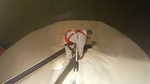 Ein Mitarbeiter in weißem Ganzkörperanzug, Haarnetz und einem roten Geschirr, das an dem ein Seil befestigt ist, steht mittig in einem Tank auf weißem Pulver. Der Mitarbeiter hält mit beiden Händen ein Rohr womit es das Pulver vermutlich aufsaugt, links neben ihm ist ein schwarze Brett um aus dem Tank ein- oder auszusteigen.