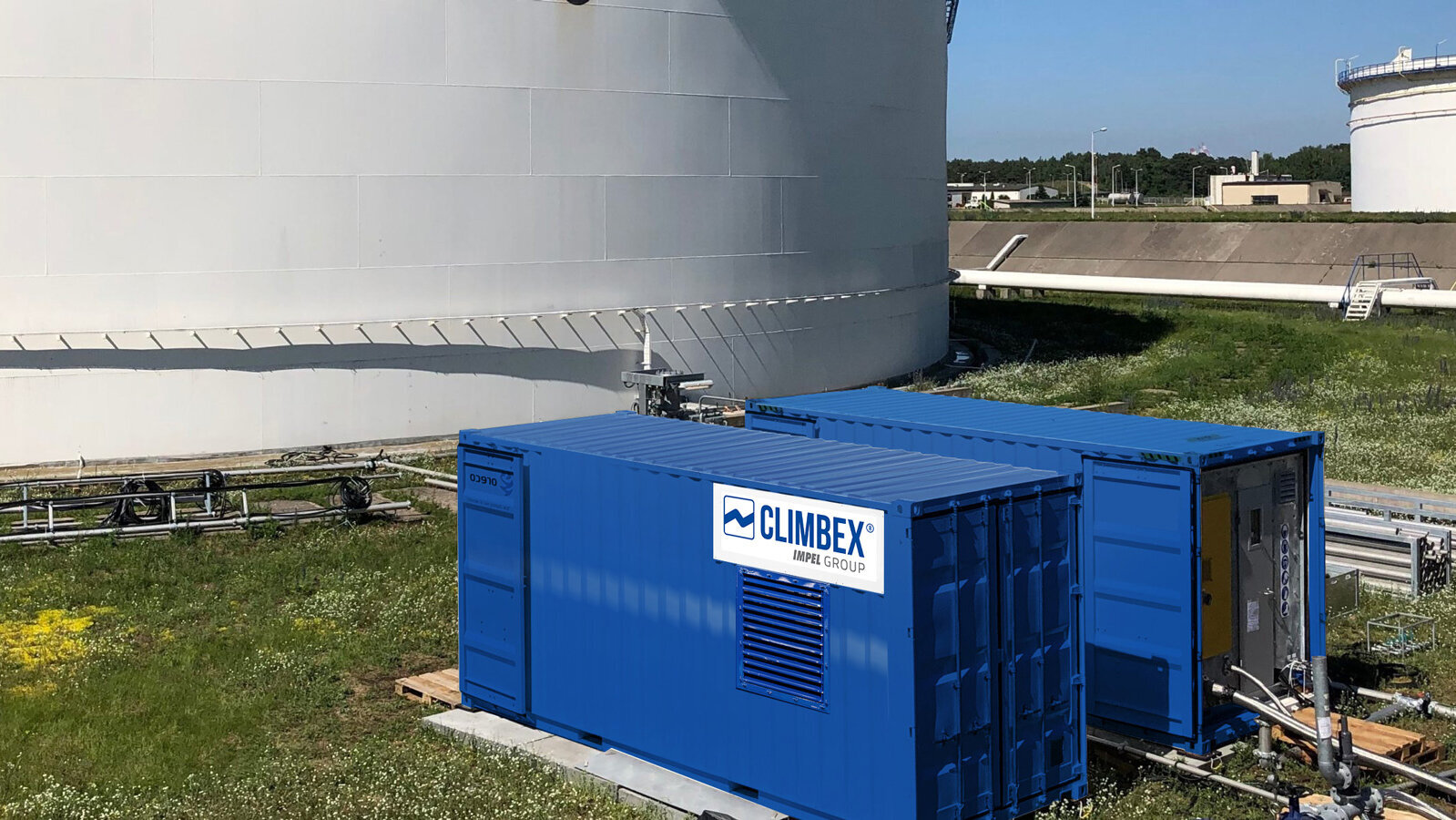 Aufnahme des Inneren eines Tanks, aufgenommen im Tank selbst. Der Boden ist Beige oder Cremefarben, es ragen viele Rohre, oben abgeklebt mit einer schwarzen Folie, aus dem Boden. Im Hintergrund ist ein Verteiler mit weiteren Rohren zu sehen, diese führen aus teilweise aus dem Bild heraus oder verbinden sich mit Ventilen aus dem Boden, die Wand des Tanks ist mit Rost versehen, darüber ist ein Stück freier blauer Himmel zu sehen. Links im Bild befindet sich eine Leiter die aus dem Tanks herausführt und rechts läuft ein Arbeiter den Tankrand entlang.