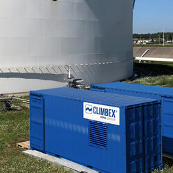 Aufnahme des Inneren eines Tanks, aufgenommen im Tank selbst. Der Boden ist Beige oder Cremefarben, es ragen viele Rohre, oben abgeklebt mit einer schwarzen Folie, aus dem Boden. Im Hintergrund ist ein Verteiler mit weiteren Rohren zu sehen, diese führen aus teilweise aus dem Bild heraus oder verbinden sich mit Ventilen aus dem Boden, die Wand des Tanks ist mit Rost versehen, darüber ist ein Stück freier blauer Himmel zu sehen. Links im Bild befindet sich eine Leiter die aus dem Tanks herausführt und rechts läuft ein Arbeiter den Tankrand entlang.