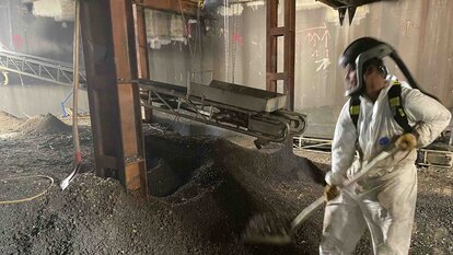 Halle mit einem Haufen an Schutt, darin stehend ein Mitarbeiter in Schutzanzug und Maske mit einer Schaufel, der Schutt auf einen Kasten auf einem Förderband auflädt. Das Förderband hängt zwischen Metallpfeilern und läuft nach hinten aus dem Bild raus.