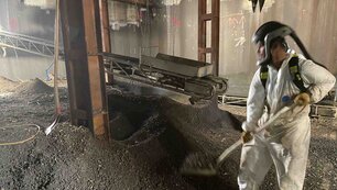 Halle mit einem Haufen an Schutt, darin stehend ein Mitarbeiter in Schutzanzug und Maske mit einer Schaufel, der Schutt auf einen Kasten auf einem Förderband auflädt. Das Förderband hängt zwischen Metallpfeilern und läuft nach hinten aus dem Bild raus.