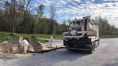 Zu sehen ist ein LKW mit Tank von hinten, an dem ein Schlauch befestigt ist, welcher von einem Arbeiter genutzt wird um eine Sand ähnlichen Berg aufzusaugen. Ein anderer Arbeiter Kehr den Sand der neben dem Berg zusammen.