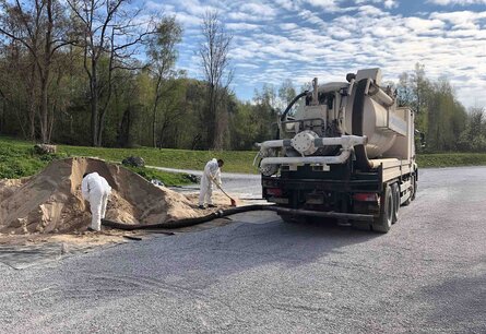 Zu sehen ist ein LKW mit Tank von hinten, an dem ein Schlauch befestigt ist, welcher von einem Arbeiter genutzt wird um eine Sand ähnlichen Berg aufzusaugen. Ein anderer Arbeiter Kehr den Sand der neben dem Berg zusammen.
