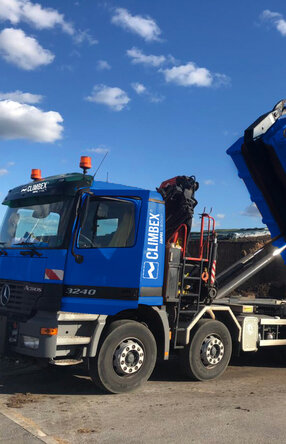 Zu sehen ist ein LKW, in der Mitte des Bildes, mit gekippter Ladefläche wie er Dreck ablädt. Hinter dem LKW steht eine Halle vor der weiterer Dreck liegt.