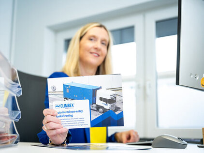 Eine Mitarbeiterin sitz am Schreibtisch, leicht unscharf, und hält vor ihrem Körper eine Climbex Broschüre in die Kamera.